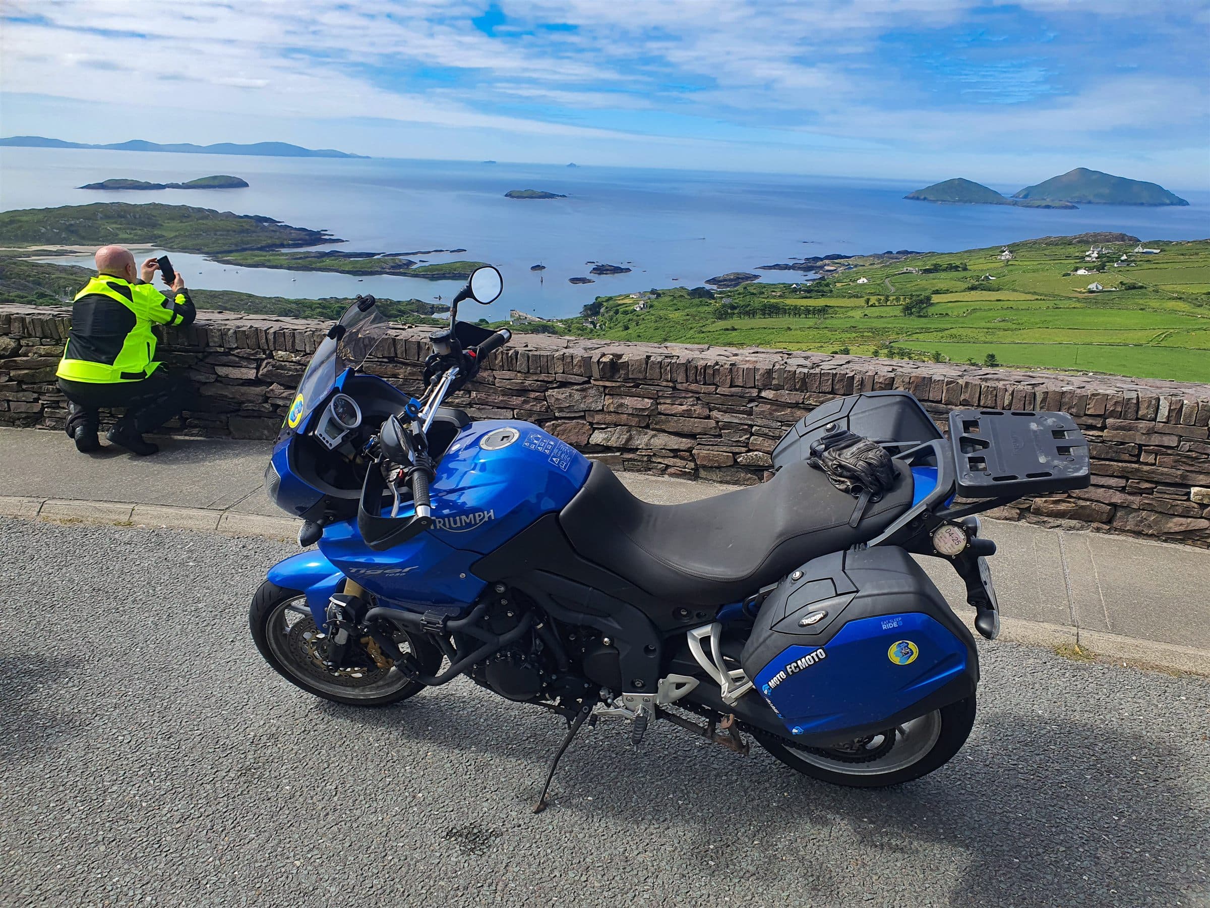 A rider photographs Derrynane Bay from the Coomakista Pass on the Ring of Kerry, his bike parked at the stone wall